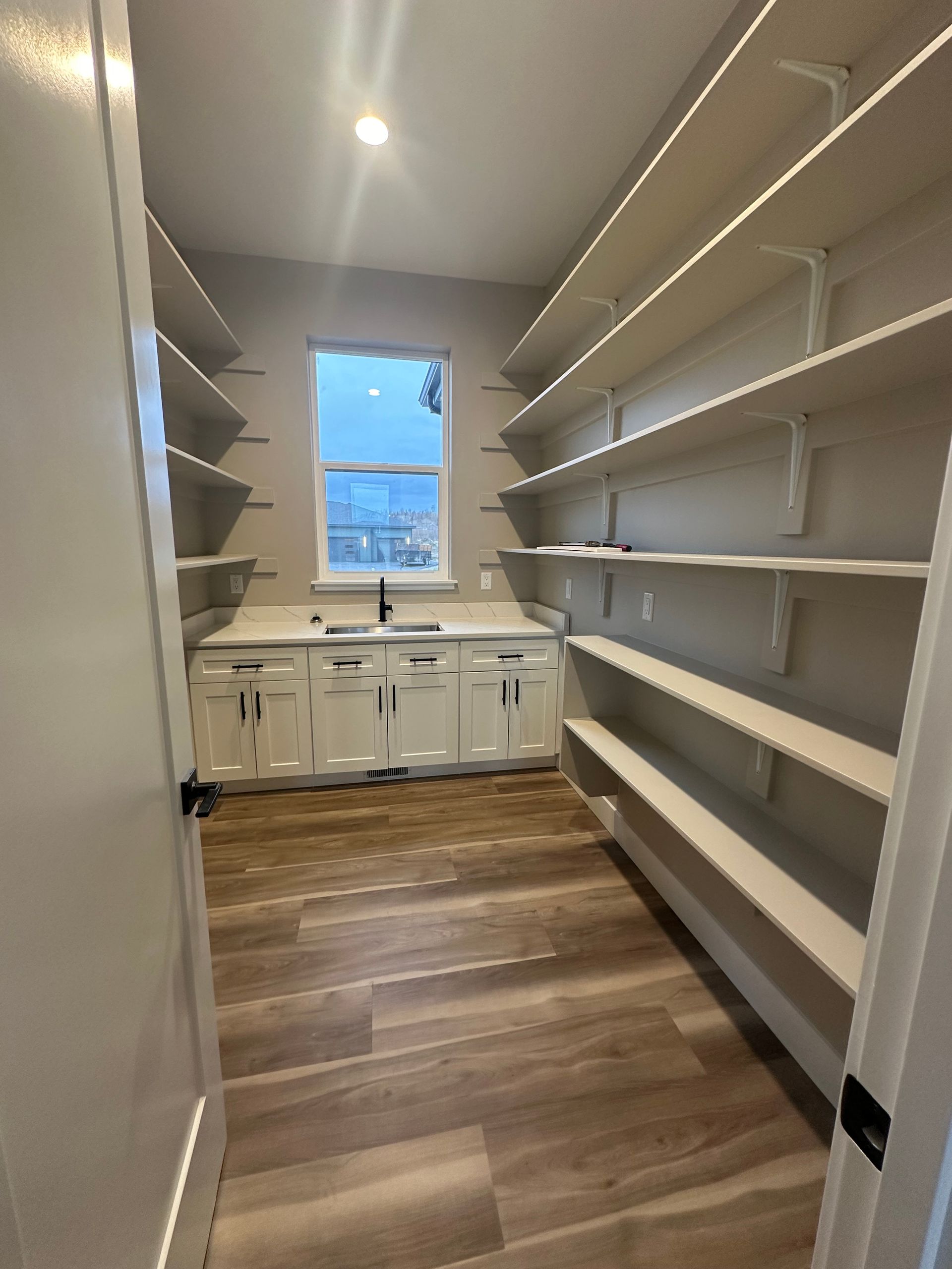 Narrow walk-in closet with white shelves and cabinets, wood floor, and a window at the far end.