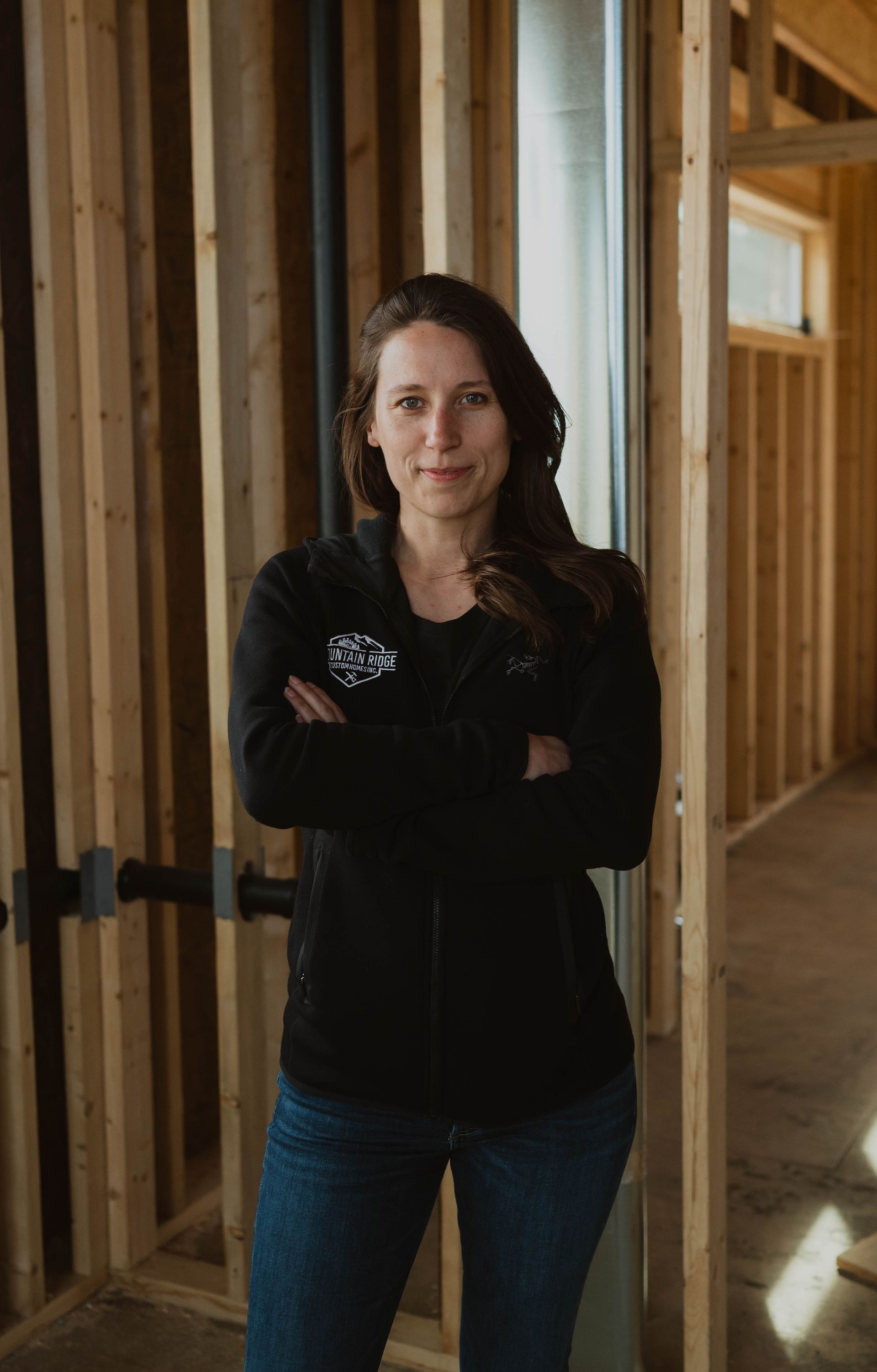 Woman standing with arms crossed in an unfinished wooden interior hallway.