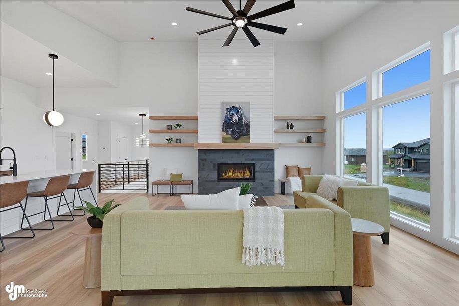 Bright modern living room with pale green sofa, fireplace, wood shelves, and large windows overlooking a yard