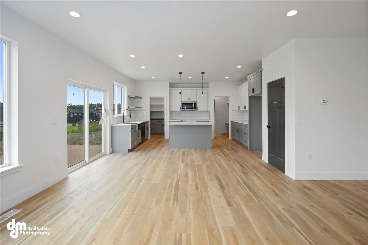 Bright, empty modern kitchen and living area with light wood floors, white walls, and large windows.