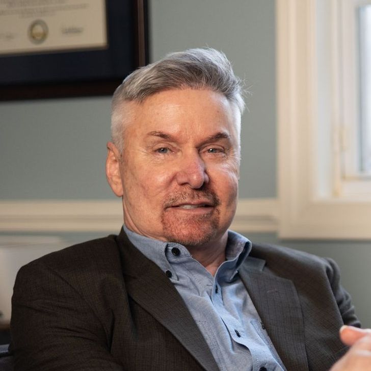 Man in blazer and blue shirt smiles, seated indoors.