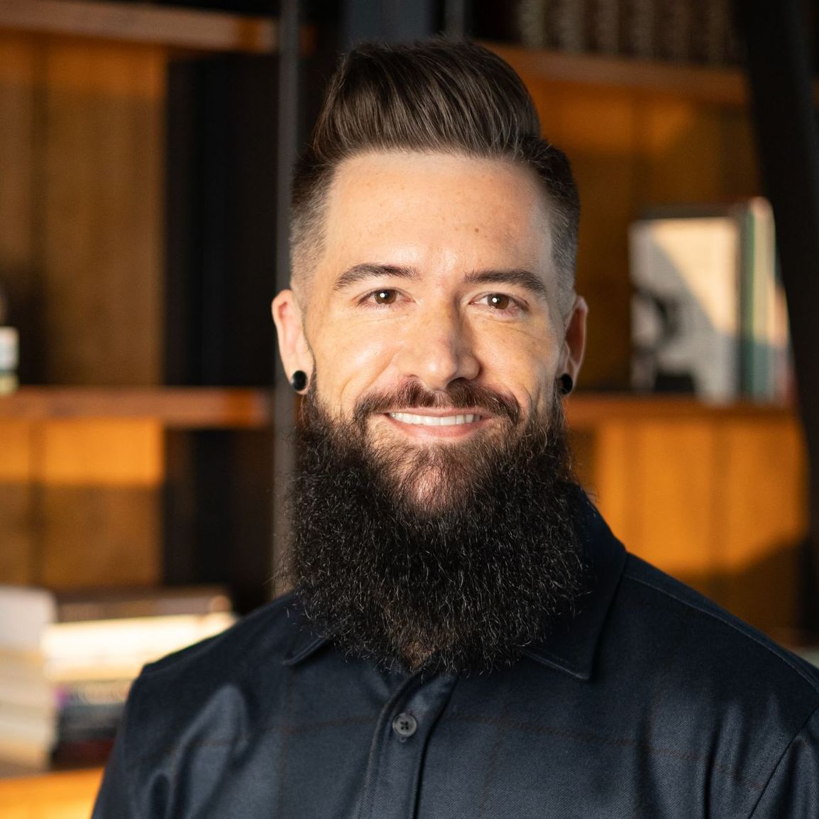 Man with dark hair and full beard smiling, wearing dark shirt; blurred bookshelf background.