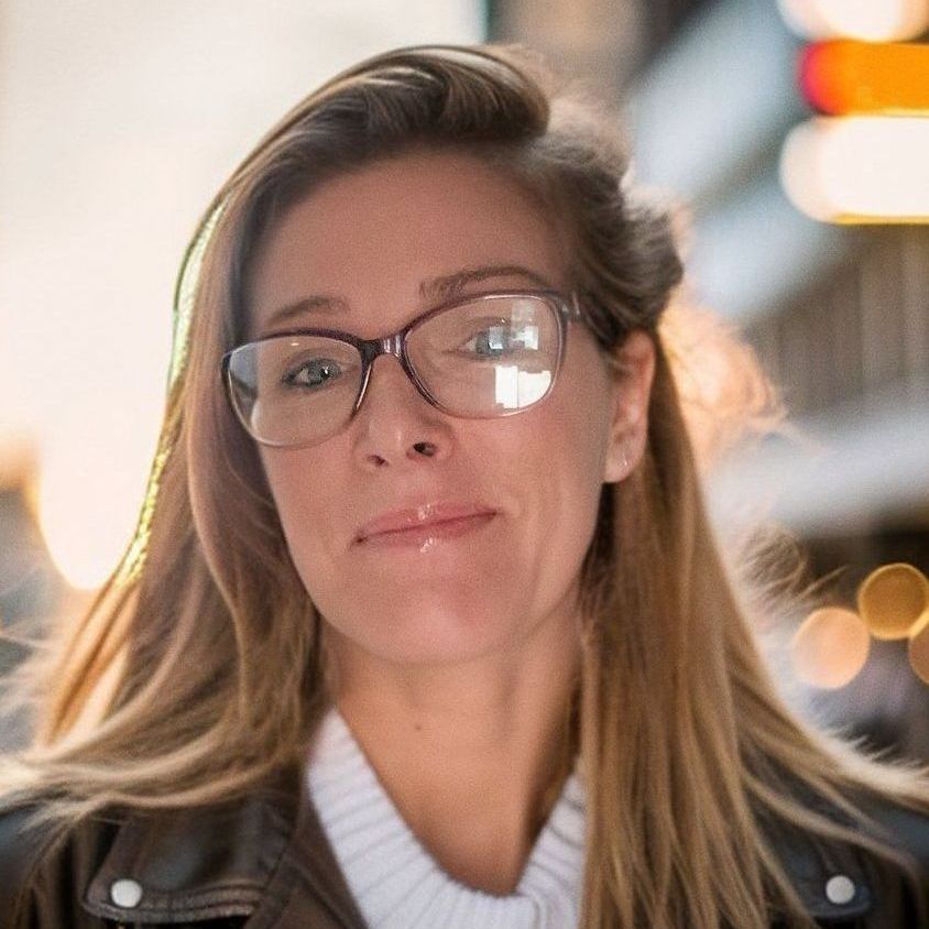 Woman with glasses looking at the camera, wearing a leather jacket, in an urban setting.