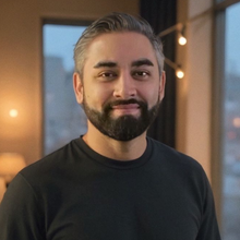Man with graying hair and beard smiles, wearing a black shirt, indoors with a window and lights.