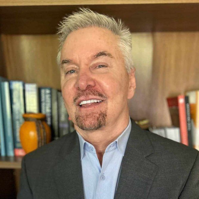 Man in blazer smiles at camera, bookshelves in background.