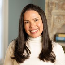 Woman with dark hair, smiling, wearing a white turtleneck sweater.
