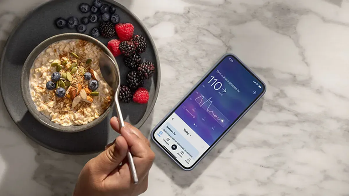 Person eating oatmeal, viewing blood glucose data on phone next to fruit.