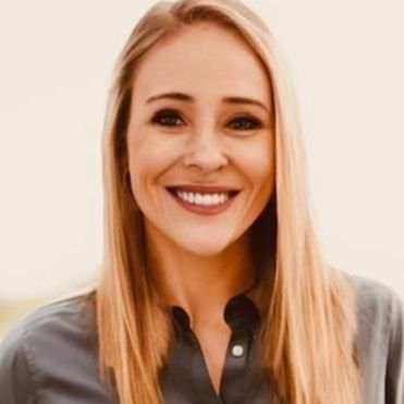 Woman with long blonde hair smiles, wearing a gray button-up shirt; outdoors, neutral background.