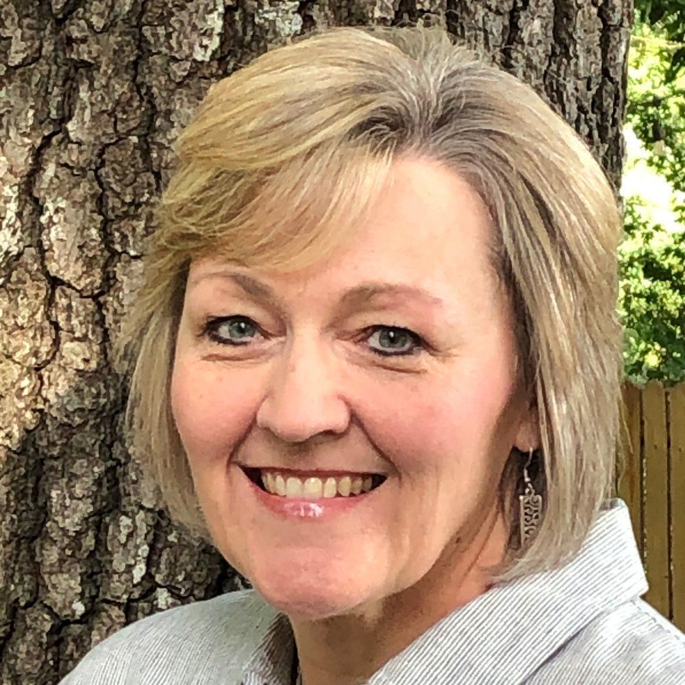 Woman with blonde hair smiling, wearing earrings, standing in front of a tree.