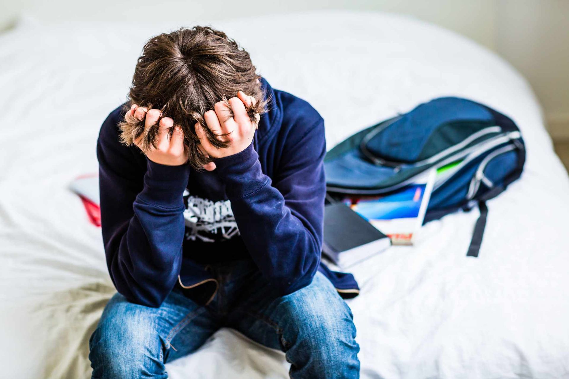 Person with head in hands, sitting on bed, backpack and books beside them.