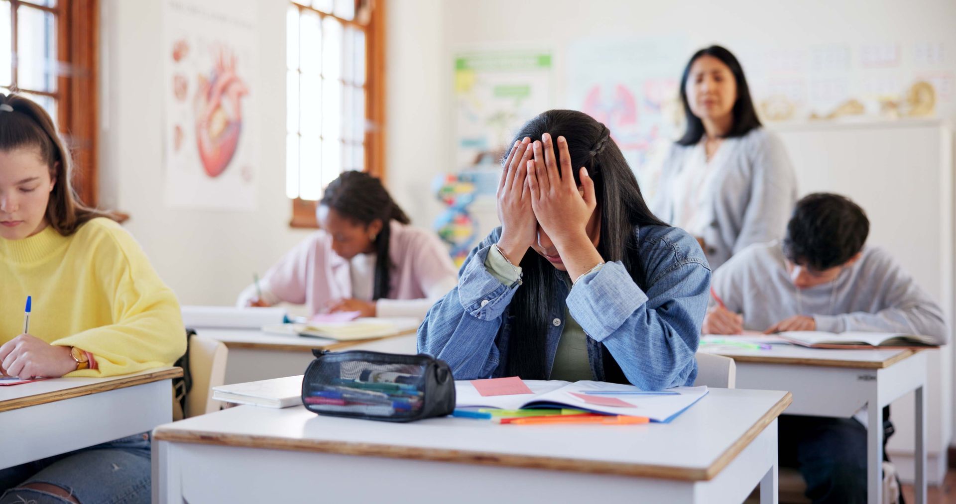 Student with head in hands at desk; classmates writing in classroom, teacher in background.