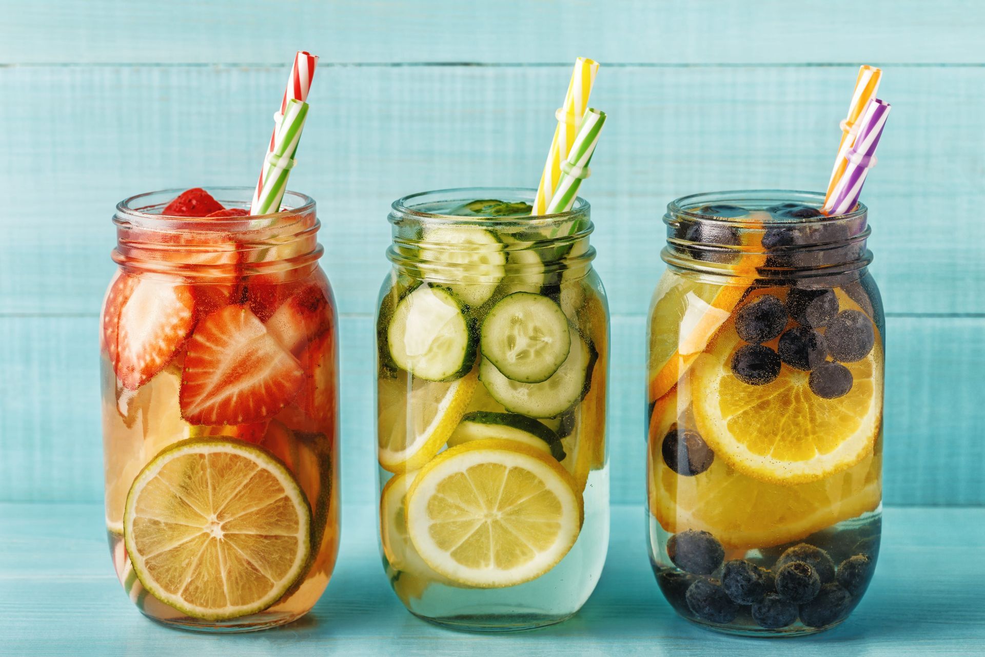 Three mason jars with fruit-infused water: strawberry, cucumber, and blueberry; on a blue wooden surface.