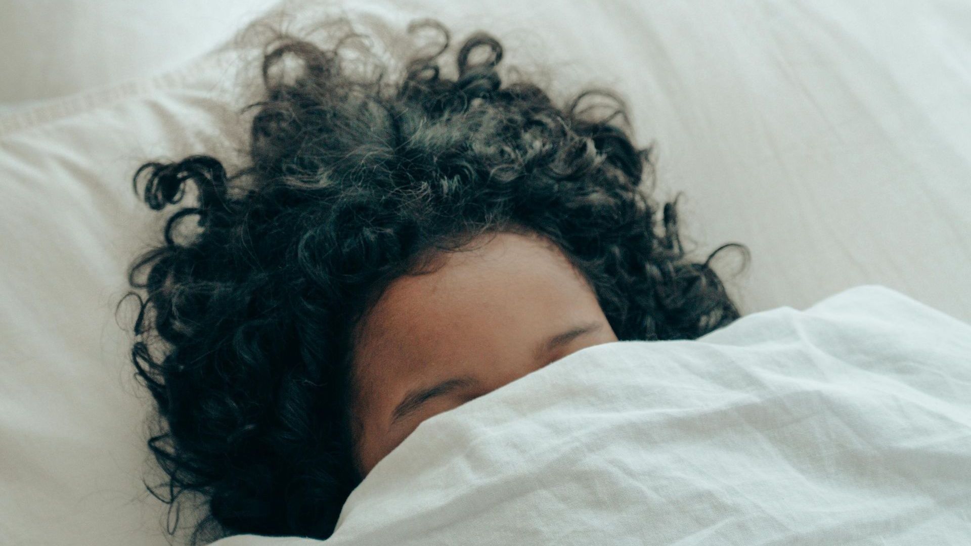 Person lying in bed, eyes covered by white blanket, curly dark hair visible.