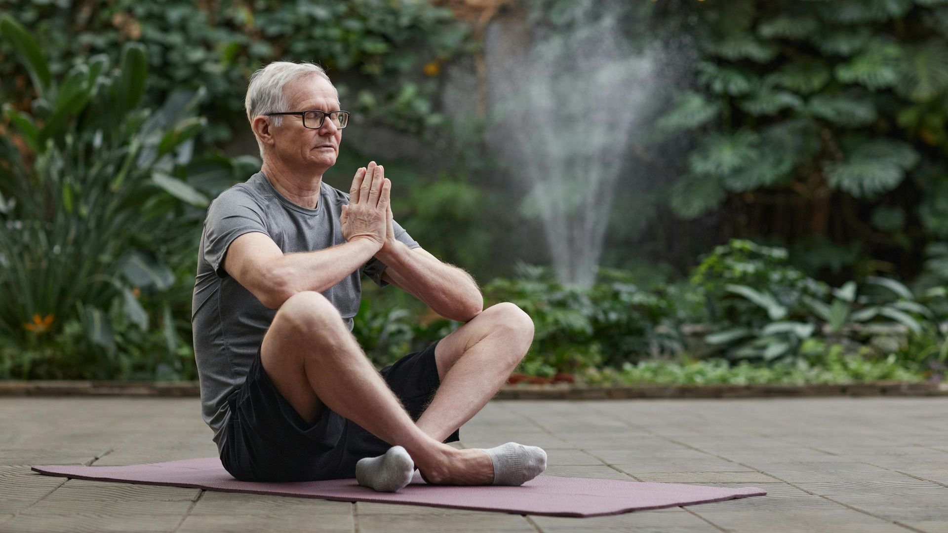 Man in gray shirt meditates on a mat outdoors, hands together, fountain in background.