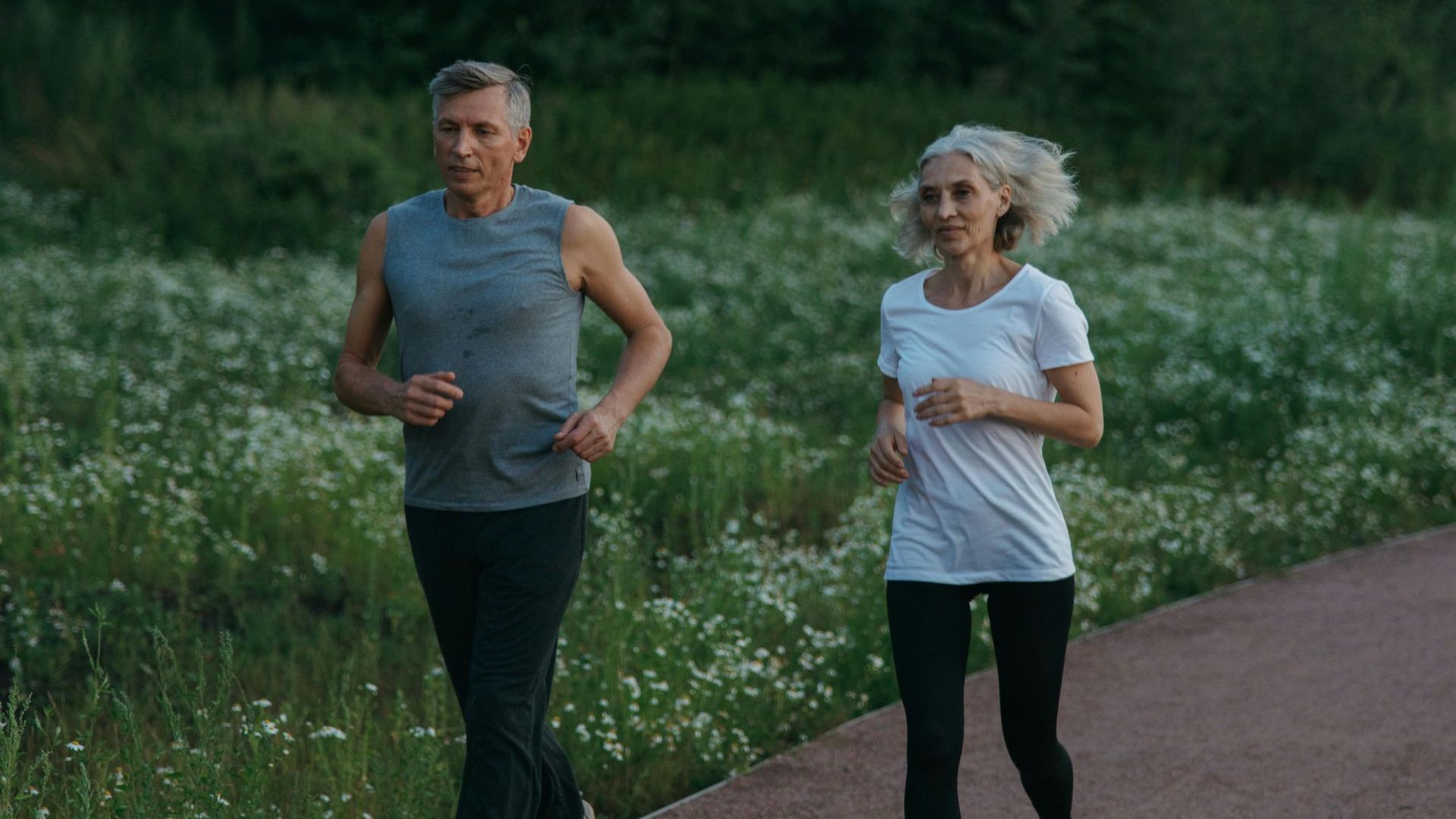 Couple jogging on a paved path in a grassy, green field. Both are wearing athletic clothing.