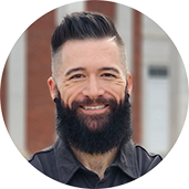Man with dark hair and full beard smiles at the camera, wearing black shirt, standing outside a brick building.