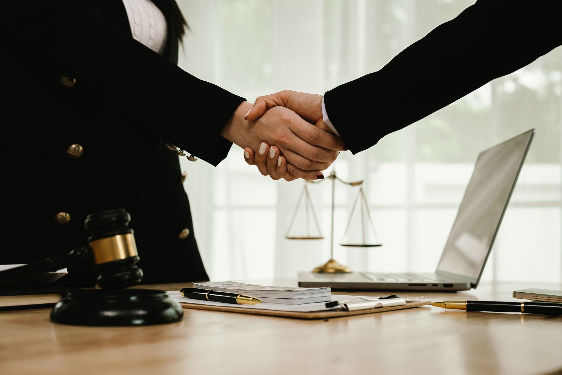 Handshake across a desk with gavel, scales, documents, and laptop in office. Handshake across a desk with gavel, scales, documents, and laptop in office.