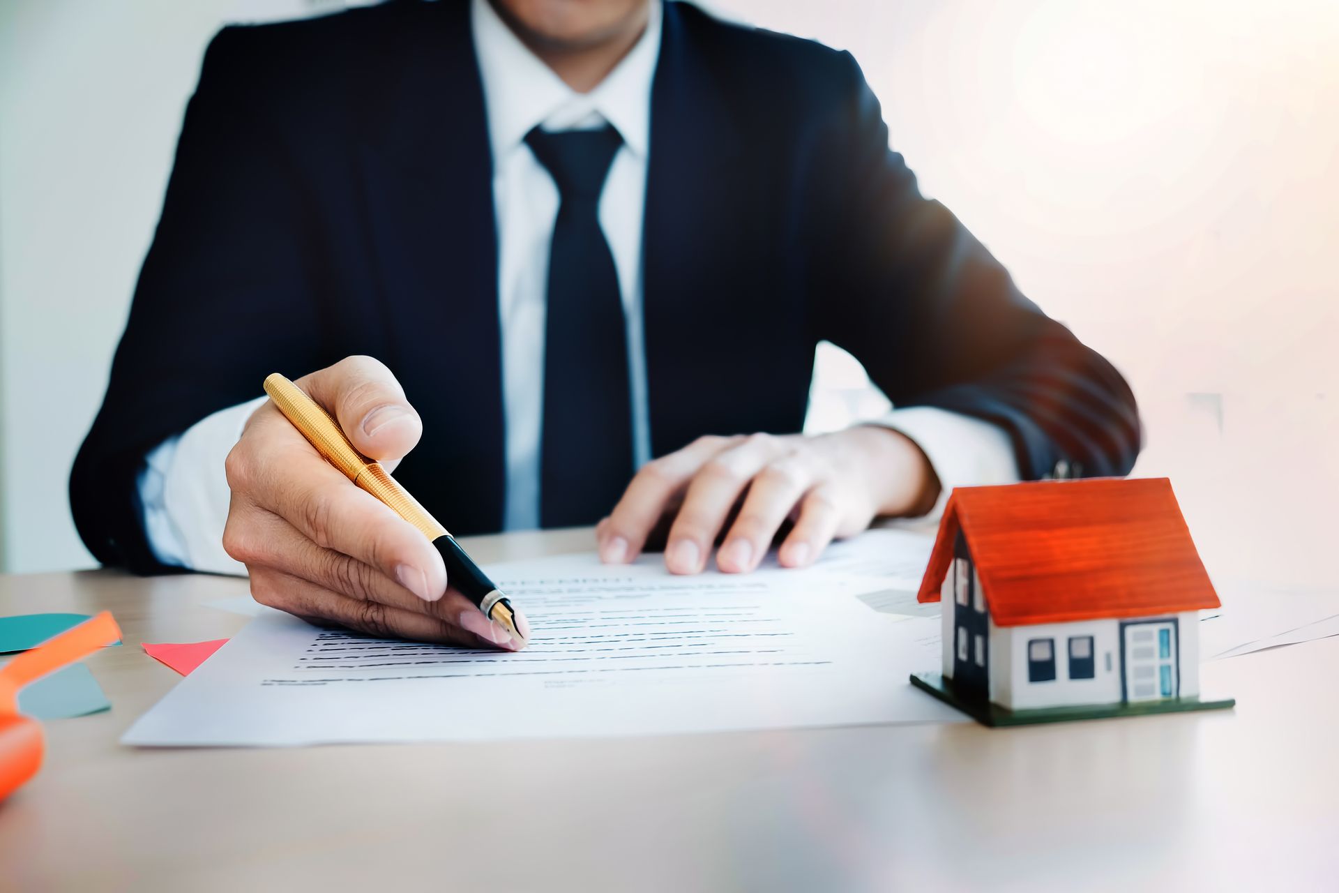 Person signing paperwork at a desk beside a small house model, suggesting a real estate.
