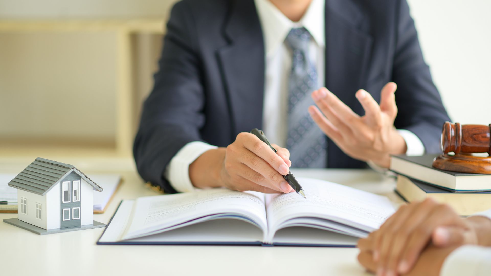 Person gesturing while writing in an open book at a desk, with a small house model nearby. Person gesturing while writing in an open book at a desk, with a small house model nearby.