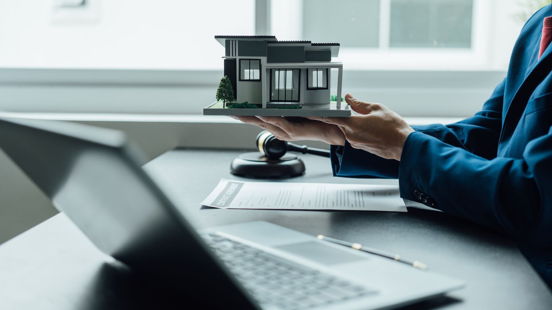 Person in suit presenting a model home over a desk with documents and laptop.