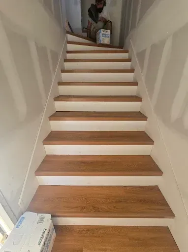 Narrow indoor staircase with white walls and wood treads, leading up to a person at the top holding a box