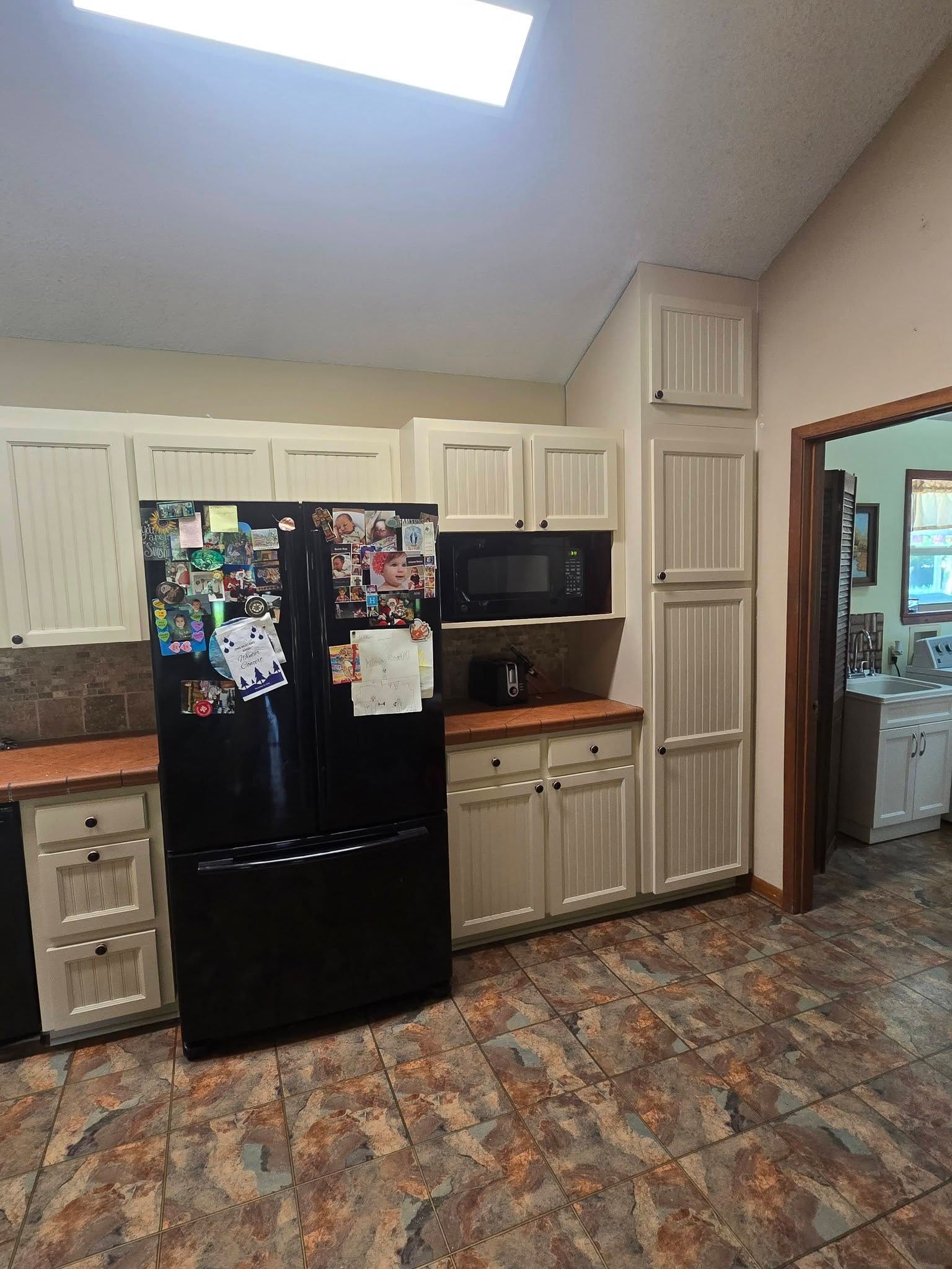 Kitchen with black fridge, white cabinets, microwave, and patterned floor under skylight.