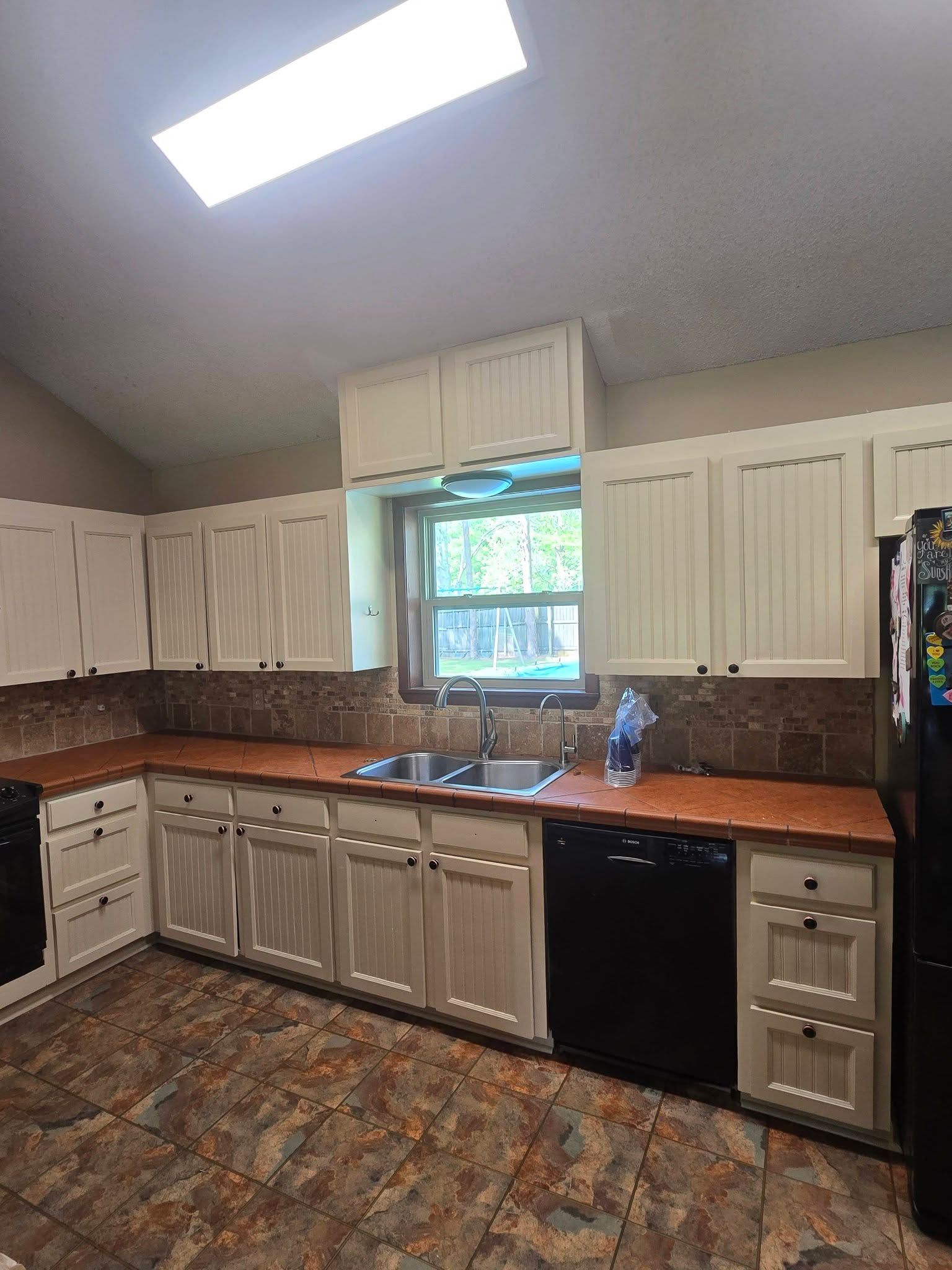 Kitchen with white cabinets, black appliances, and a skylight over a sink beneath a window.