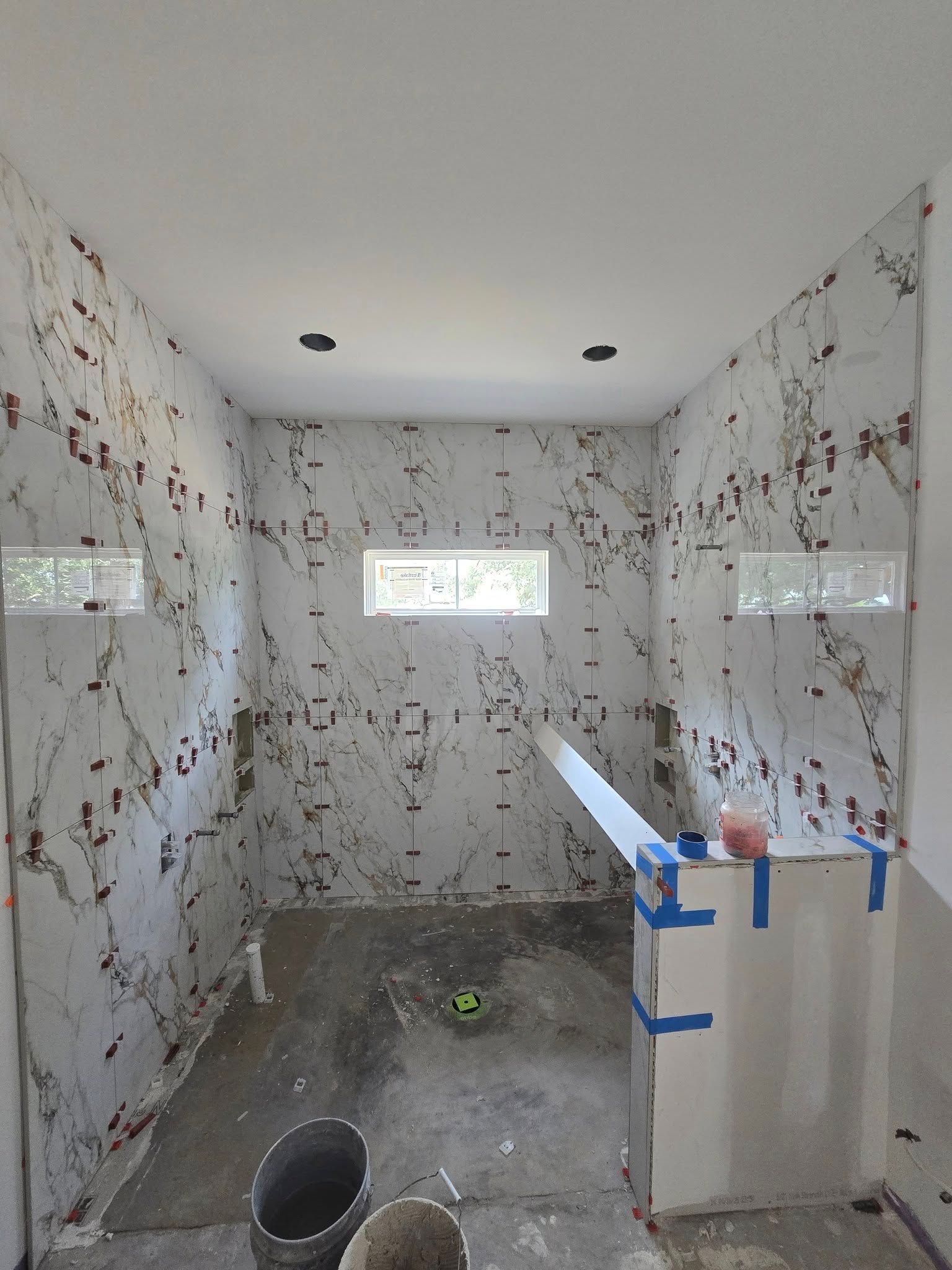 Partially tiled bathroom under construction with two small windows, unfinished floor, and a sink wrapped in blue tape