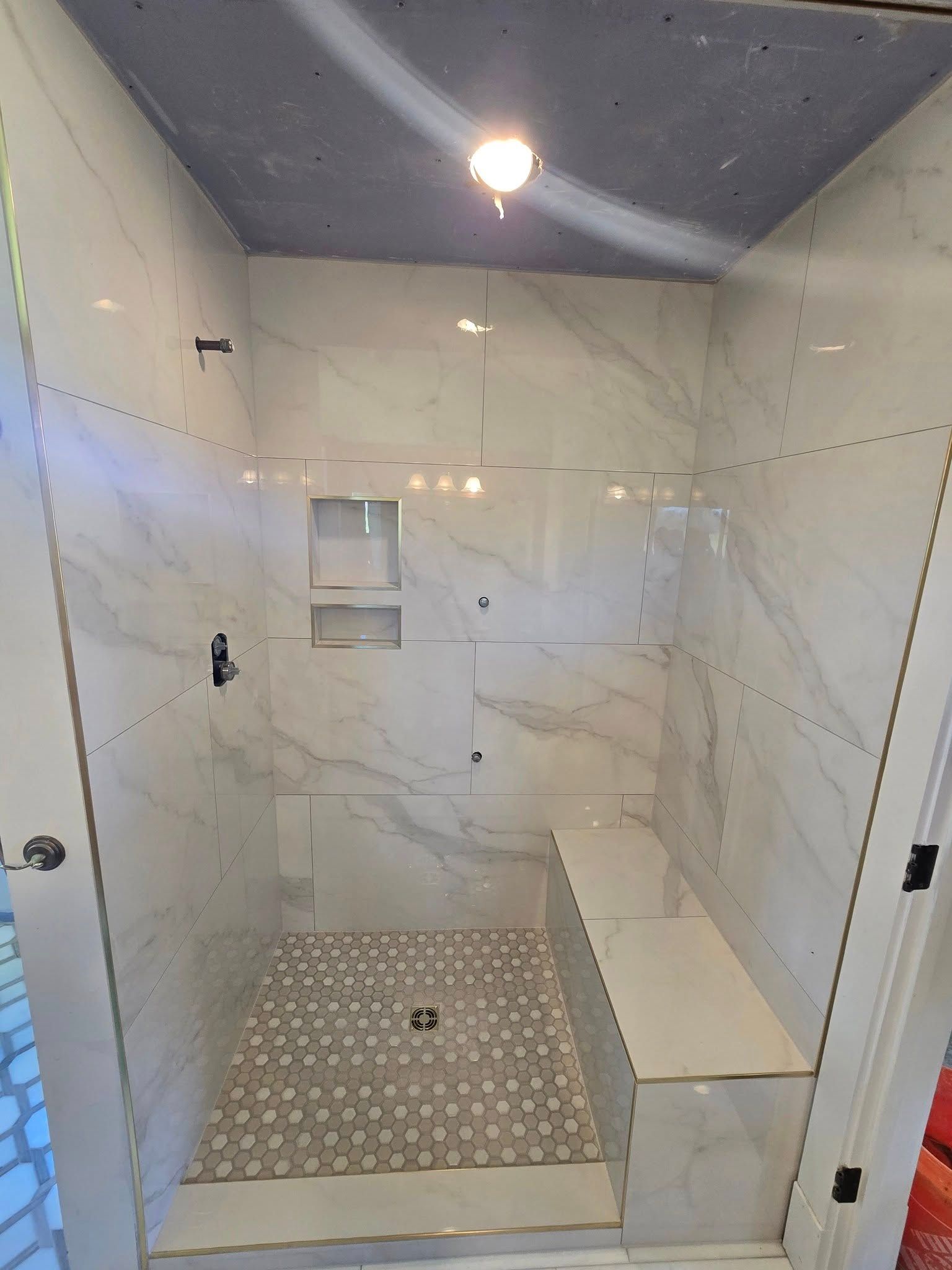 Empty tiled shower with marble-look walls, mosaic floor, built-in bench, and ceiling light.