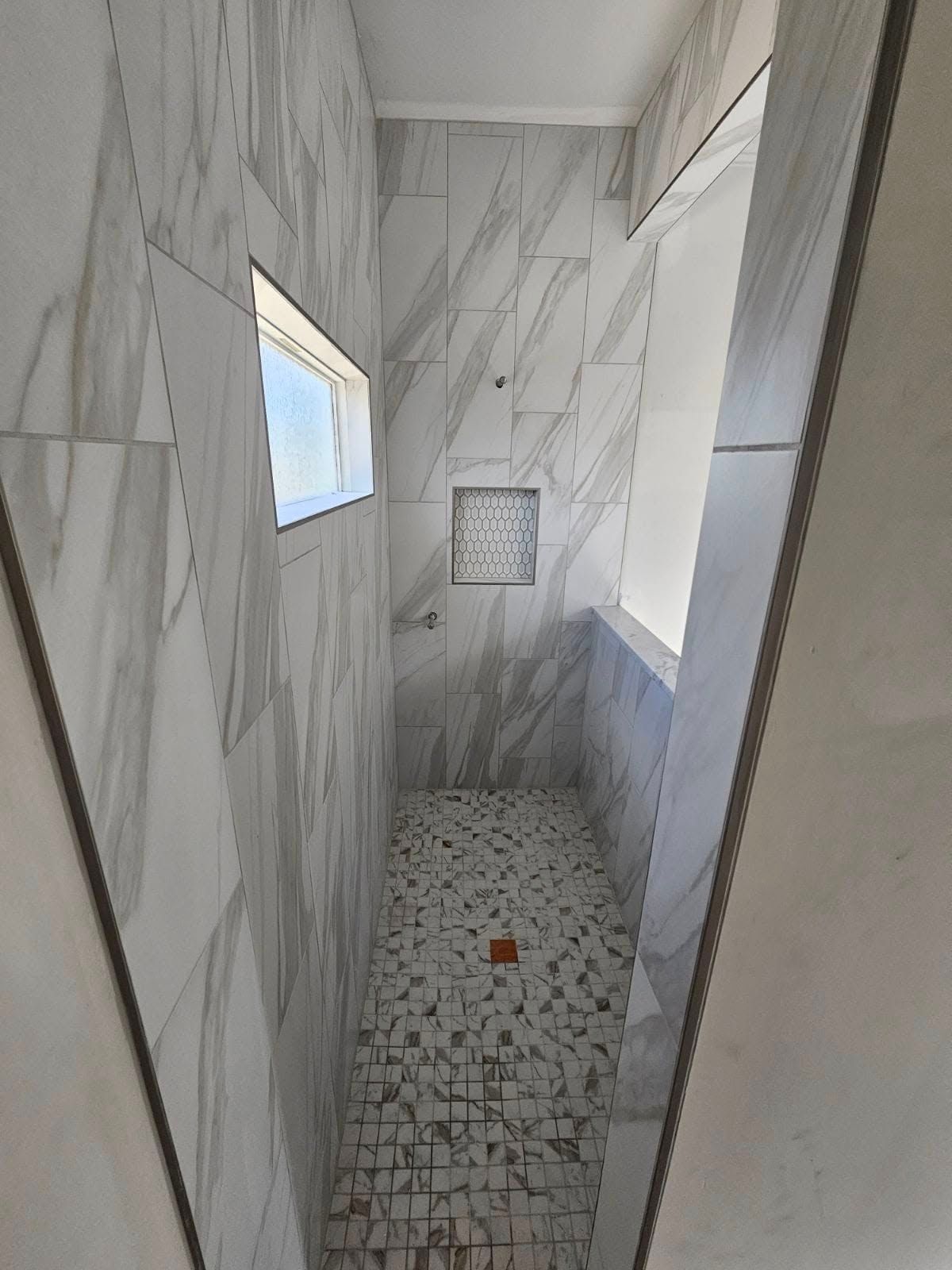 Narrow tiled shower corridor with white marble-look walls, pebble floor, and small windows.