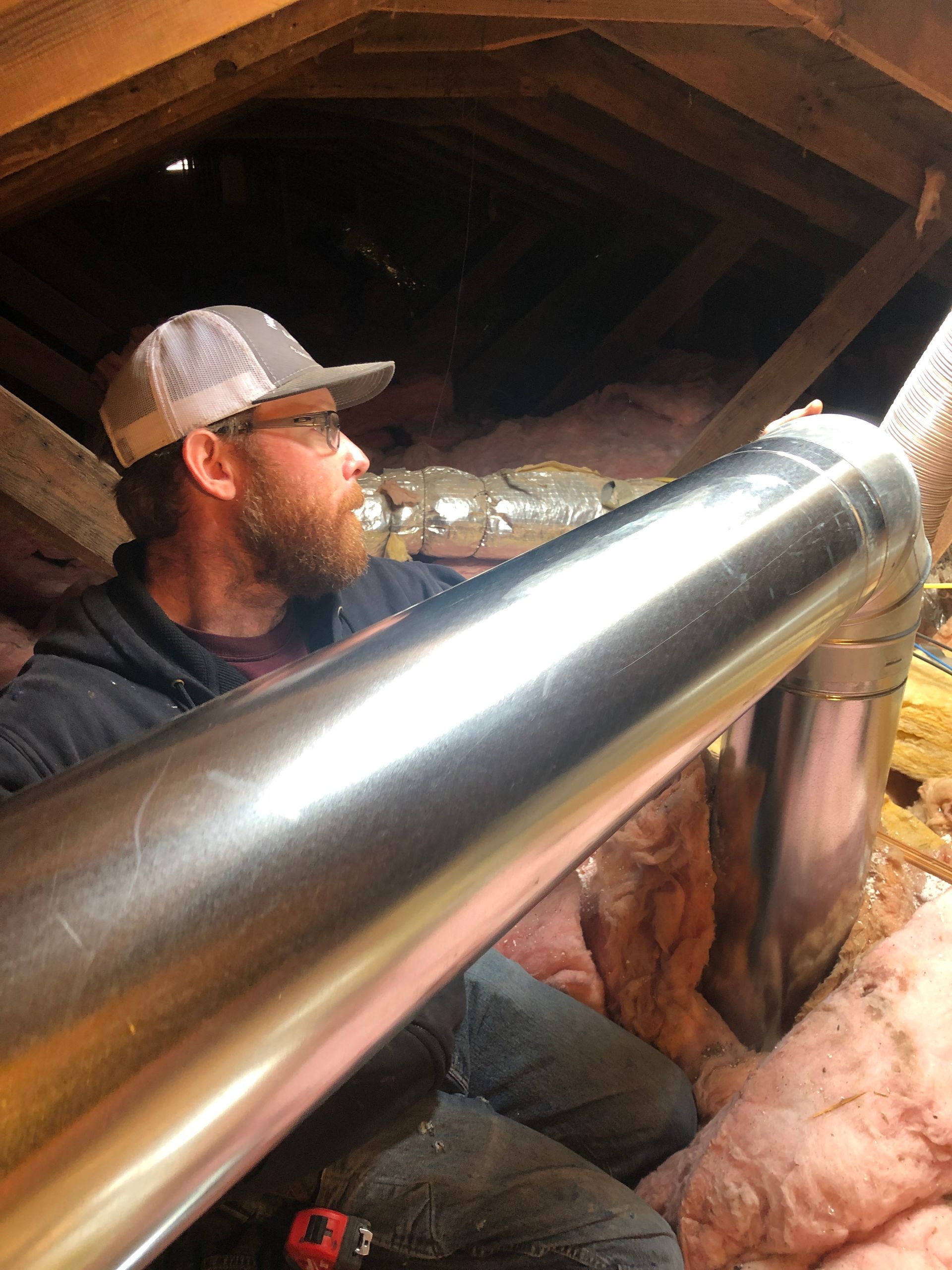 Man working in an attic with exposed wooden beams and insulated pipes.