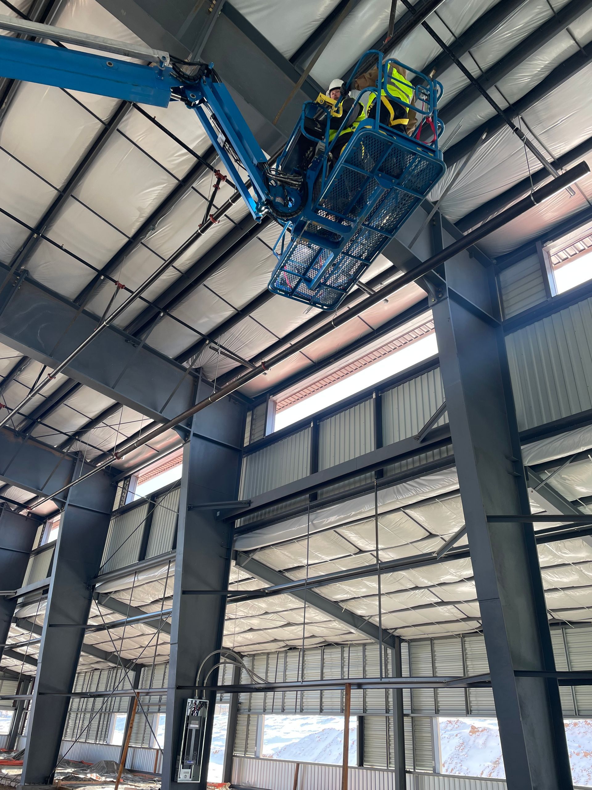 Workers in a blue boom lift inspect the ceiling inside a large industrial warehouse.