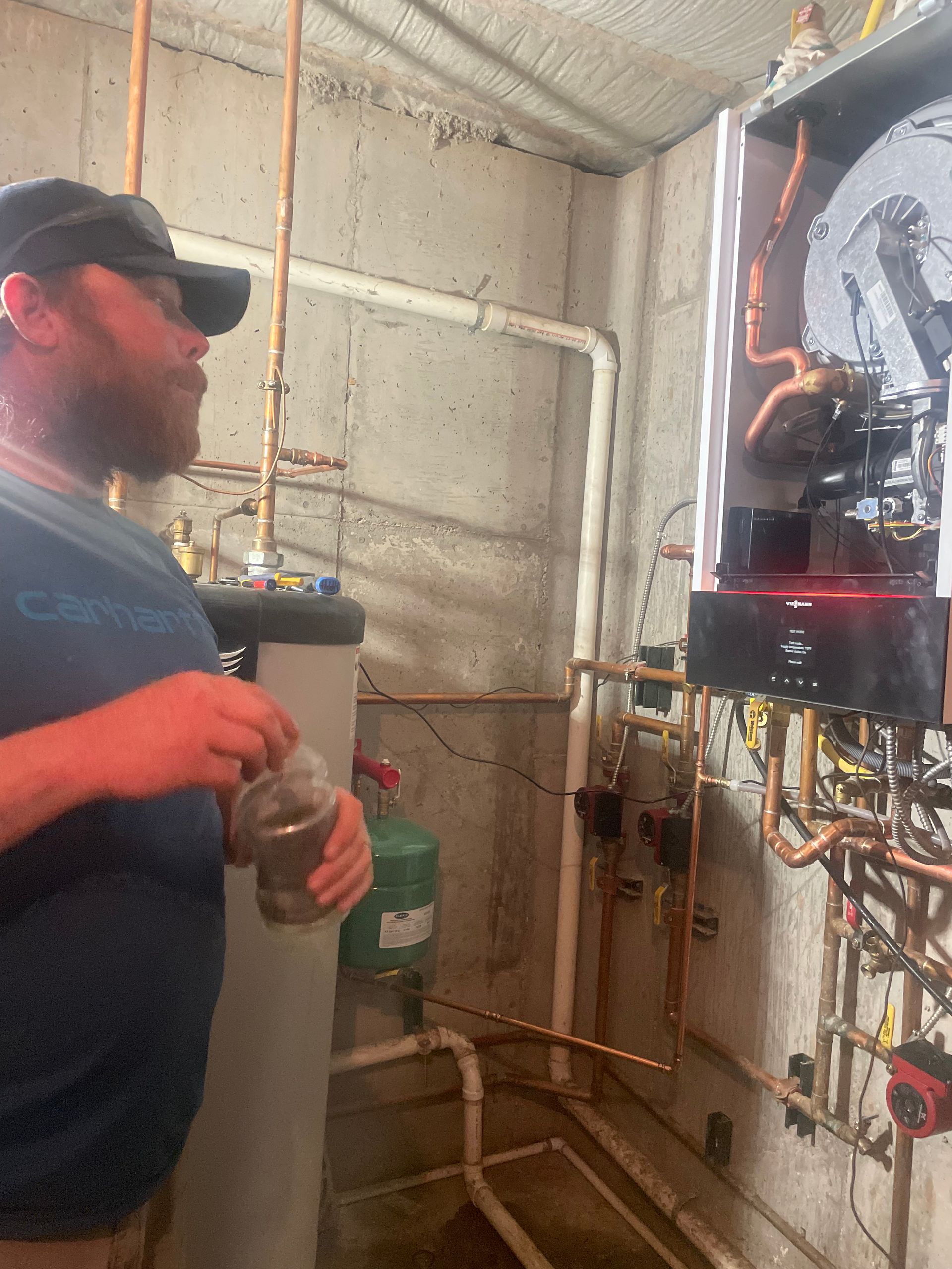 Worker inspecting electrical equipment in a utility room, holding a drink and looking at exposed wiring.