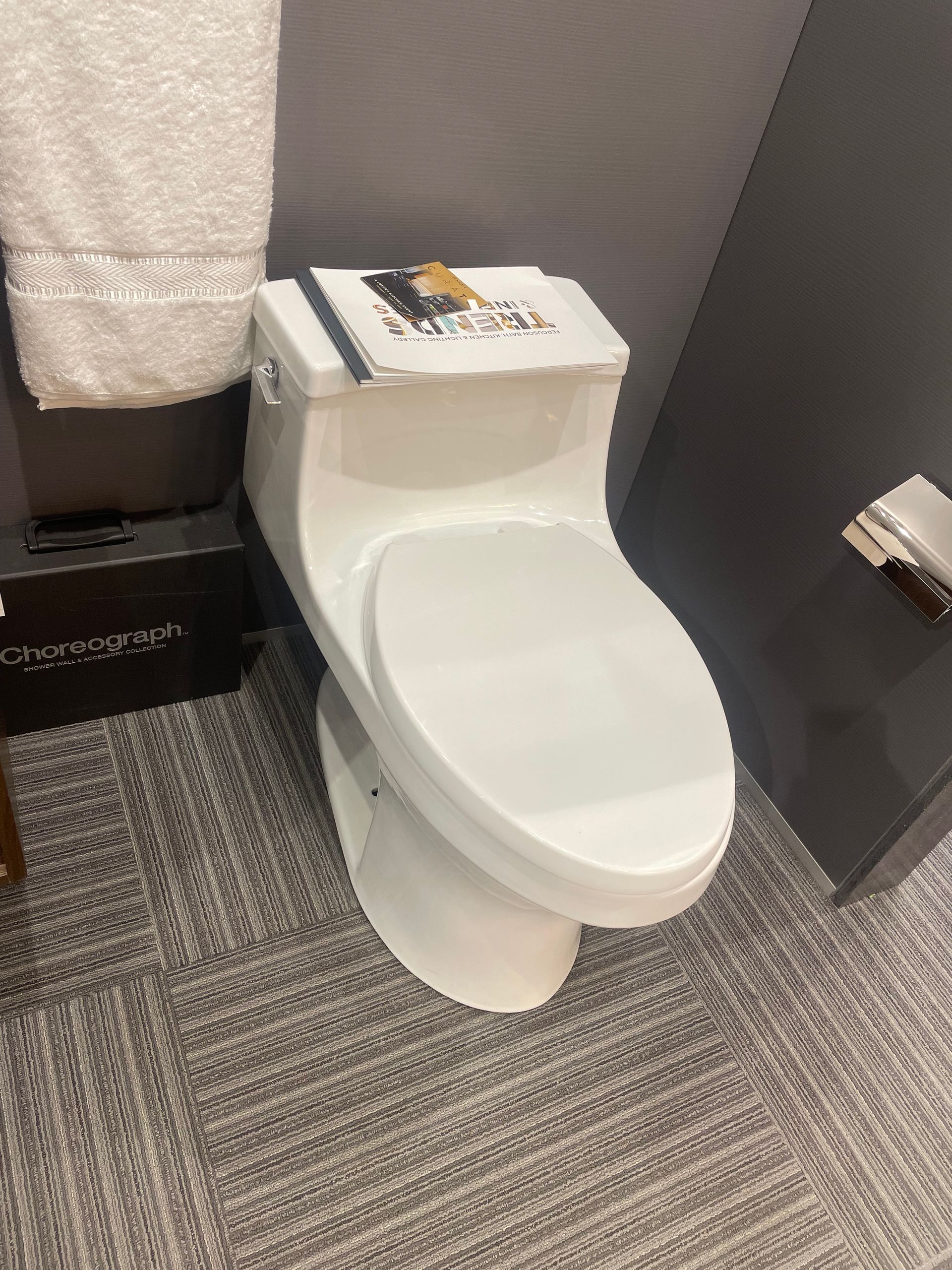 White toilet in a gray-tiled bathroom with a towel on the wall and magazines on the tank.