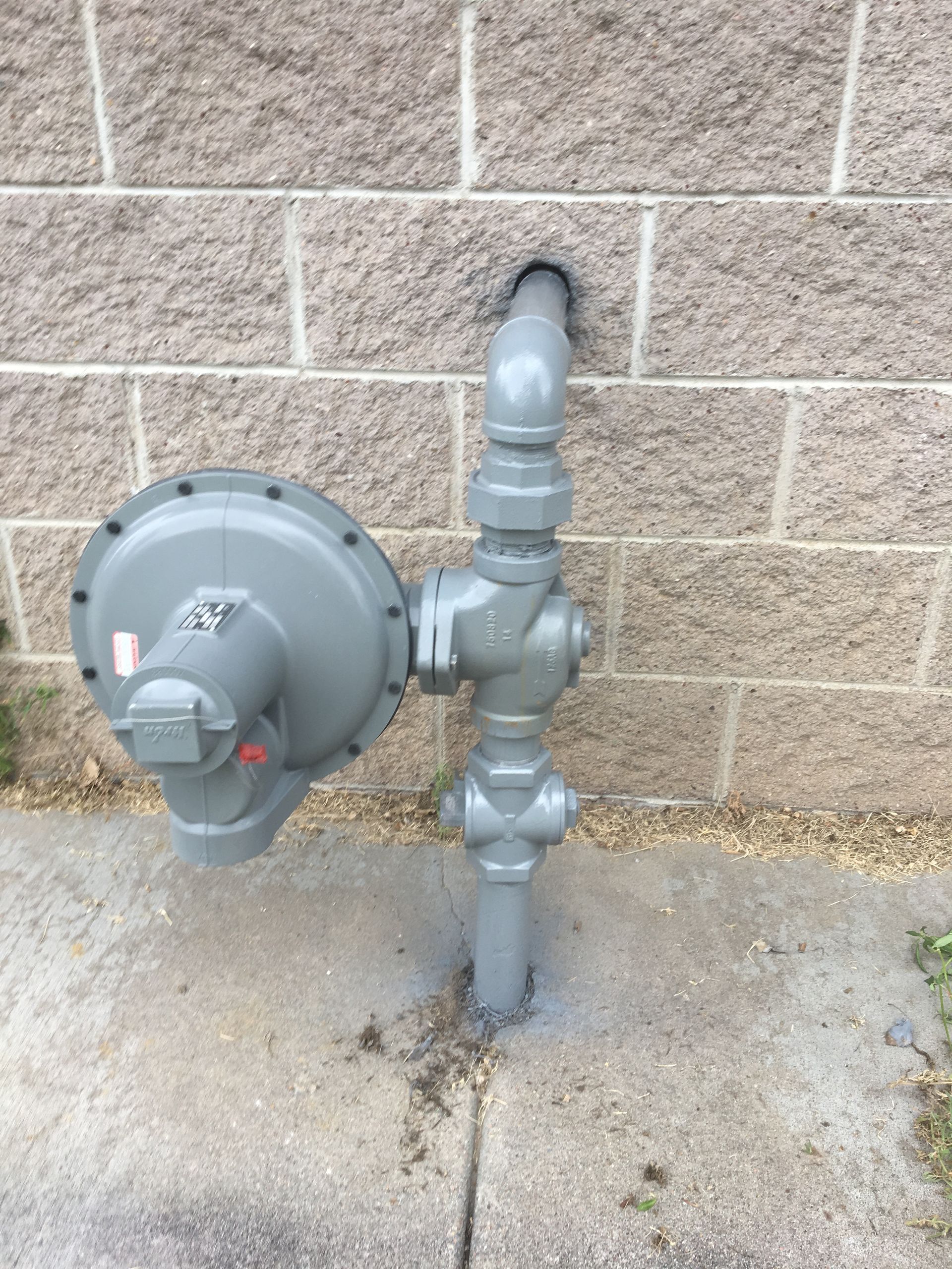 Gas meter and utility pipes mounted on a brick wall beside a concrete walkway.