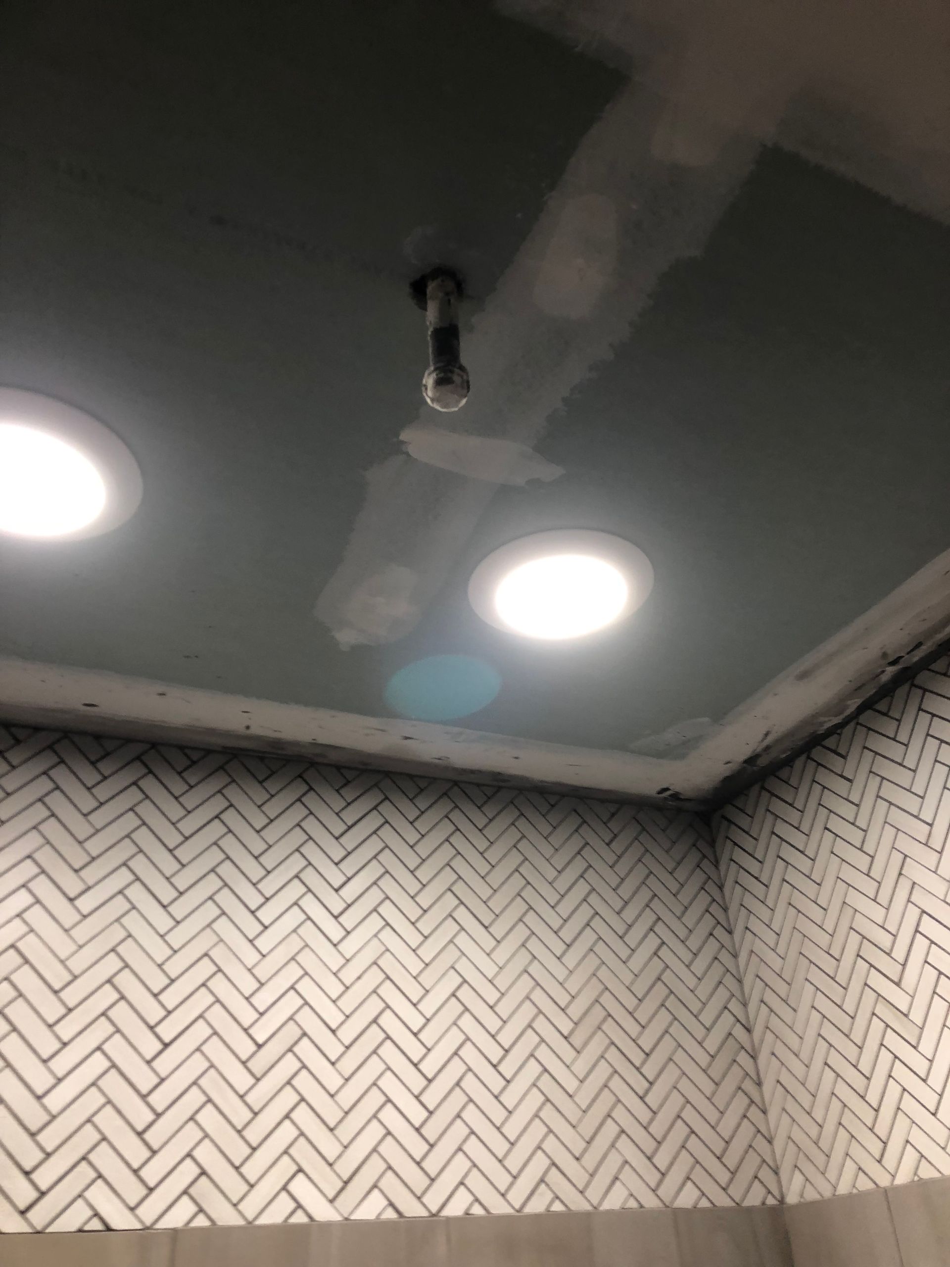 Bathroom ceiling with two recessed lights and a central exposed pipe above herringbone-patterned wall tiles