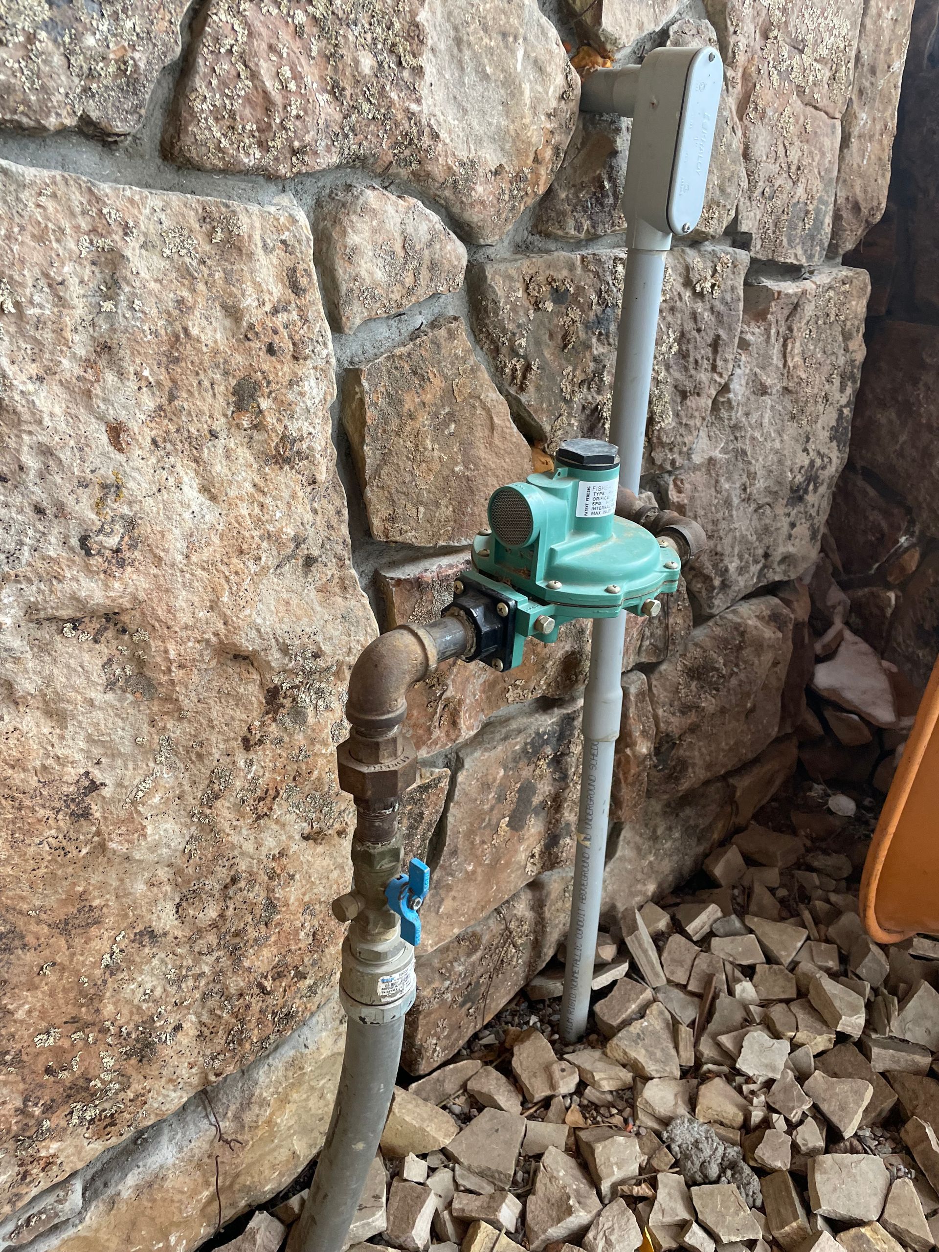 Outdoor water meter and pipe assembly mounted on a stone wall, with gravel below