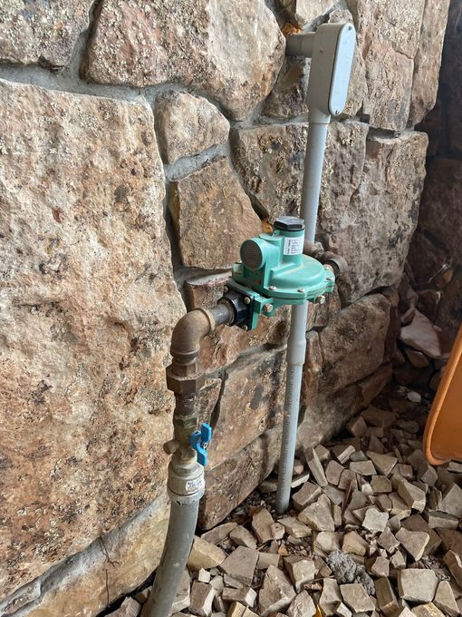 Outdoor water meter and pipe assembly mounted on a stone wall, with gravel below