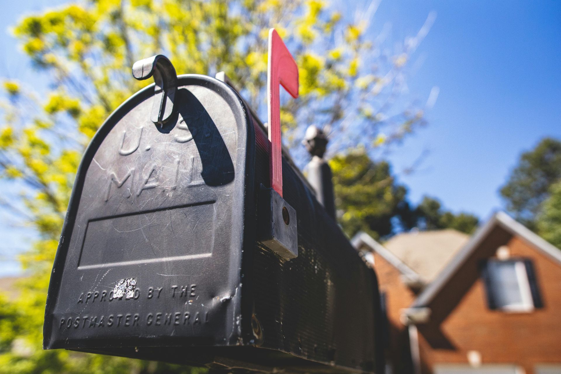 Black mailbox with the flag up, set against a blurred house and sunny sky.