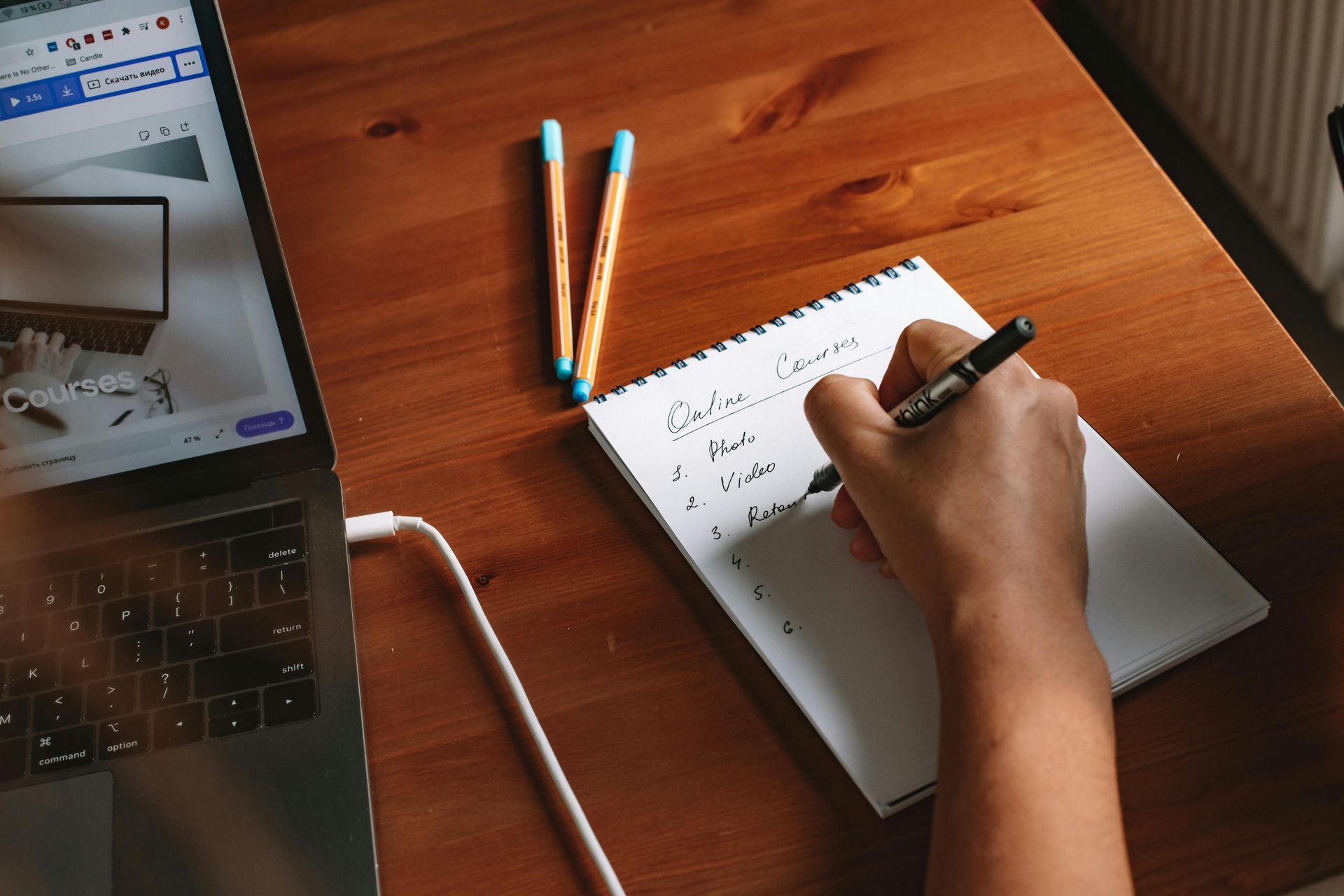 Person writing in a notebook on a wooden desk, laptop open on the left, two pens.