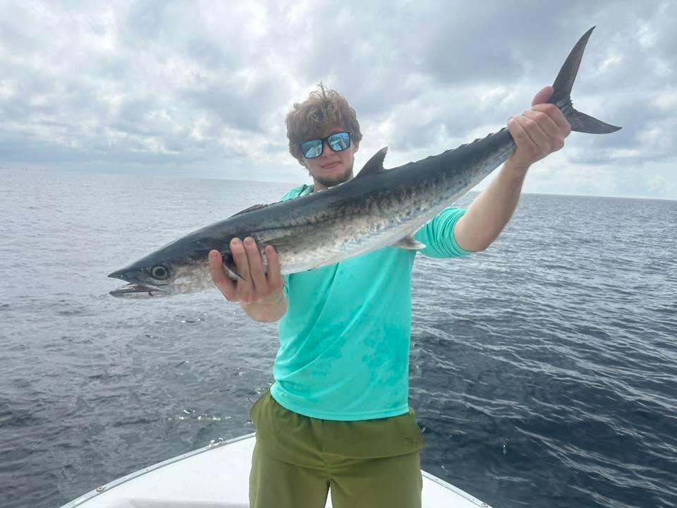 Person holding a large fish on a boat, wearing a turquoise shirt and sunglasses, with the ocean behind them.