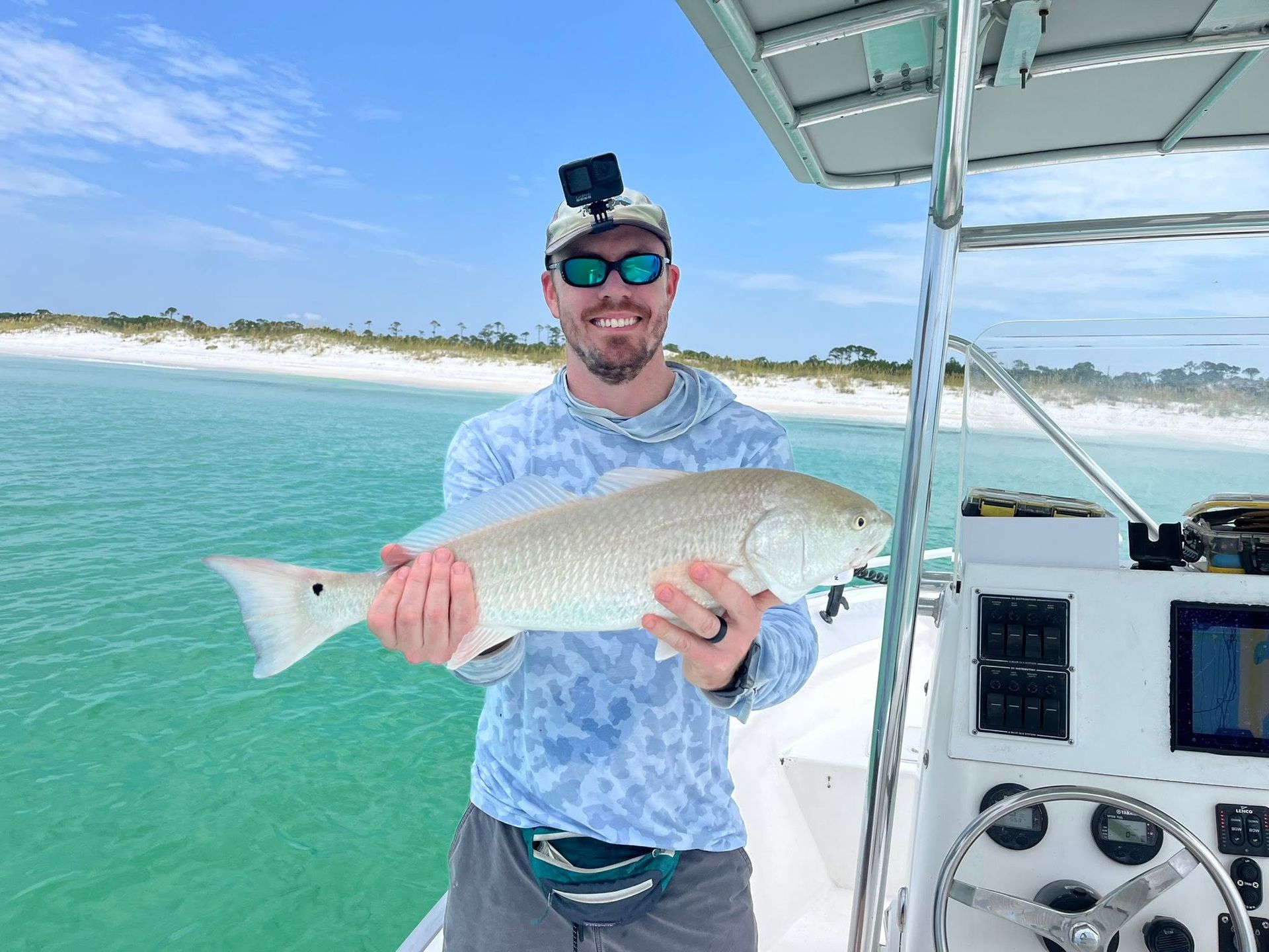 Man holding a large fish on a boat in clear green water near a sandy beach