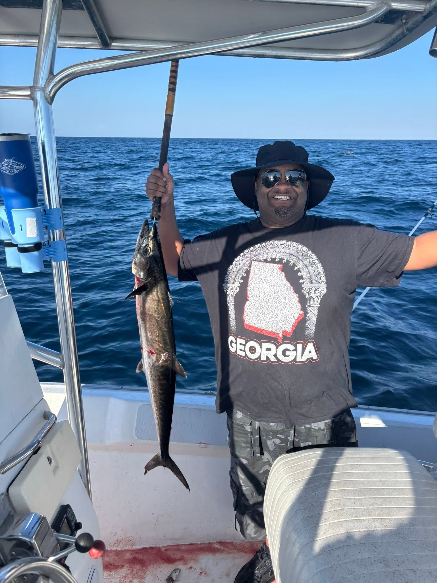 Person on a boat holding a long fish over the ocean, wearing a black hat and sunglasses.