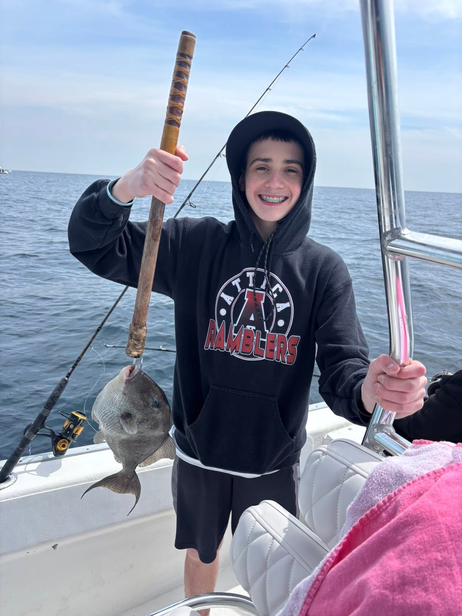 Smiling person in a hoodie on a boat, holding up a fish on a fishing rod with the ocean behind them