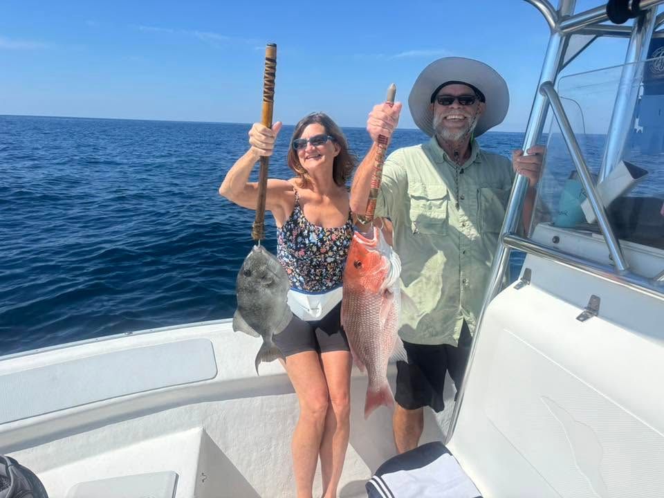 Two people on a boat holding up large fish, smiling on the open ocean