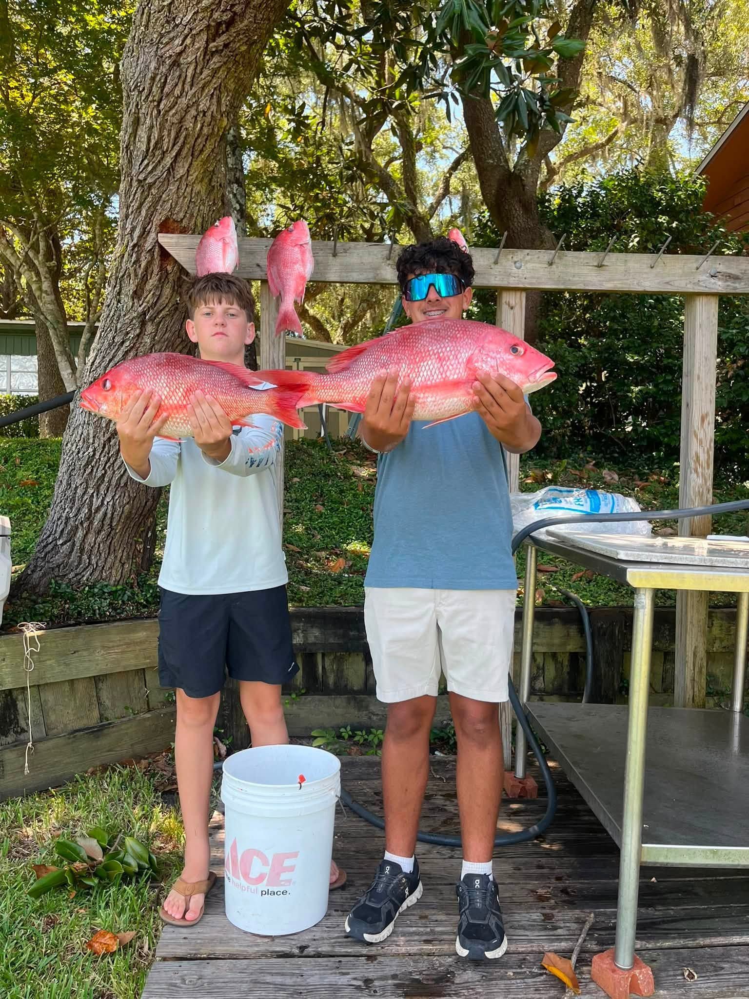 Two people outdoors holding large red-orange fish beside a white bucket and table under trees