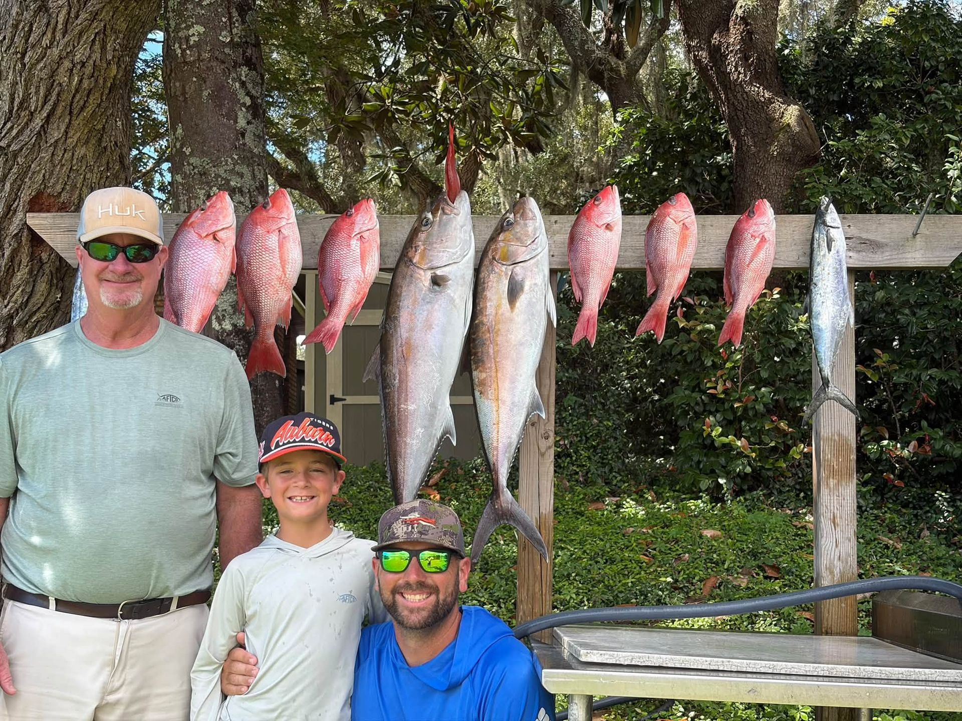 Three people posing beside a row of hanging fish outdoors near a picnic table