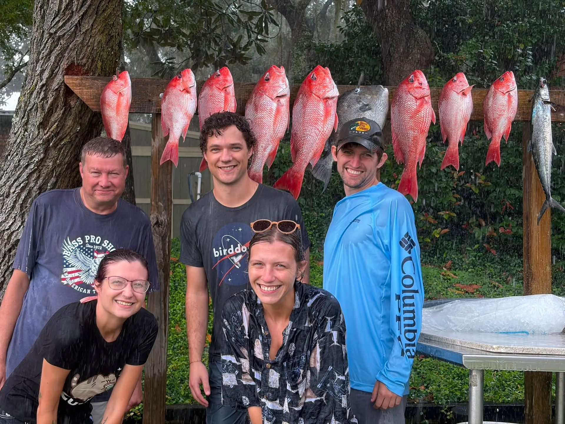 Five people pose beneath hanging red fish at an outdoor fish-cleaning station.