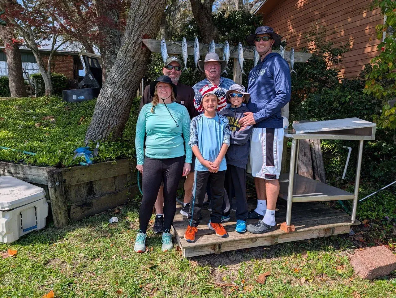 Group of six people posing outdoors on a small platform by a brick wall and large tree.