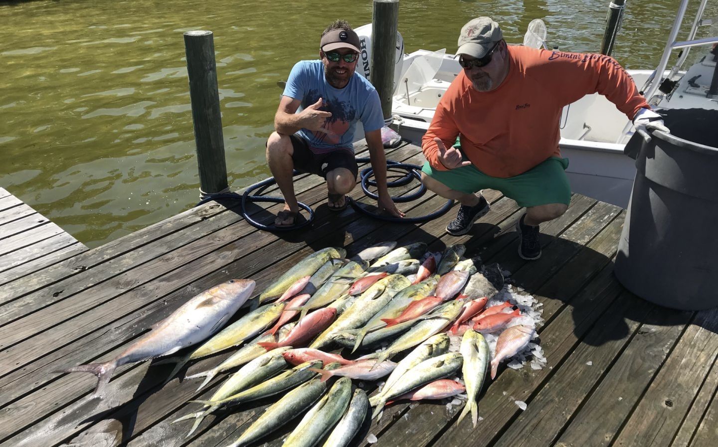 Two people kneel beside a large catch of colorful fish on a dock by the water.
